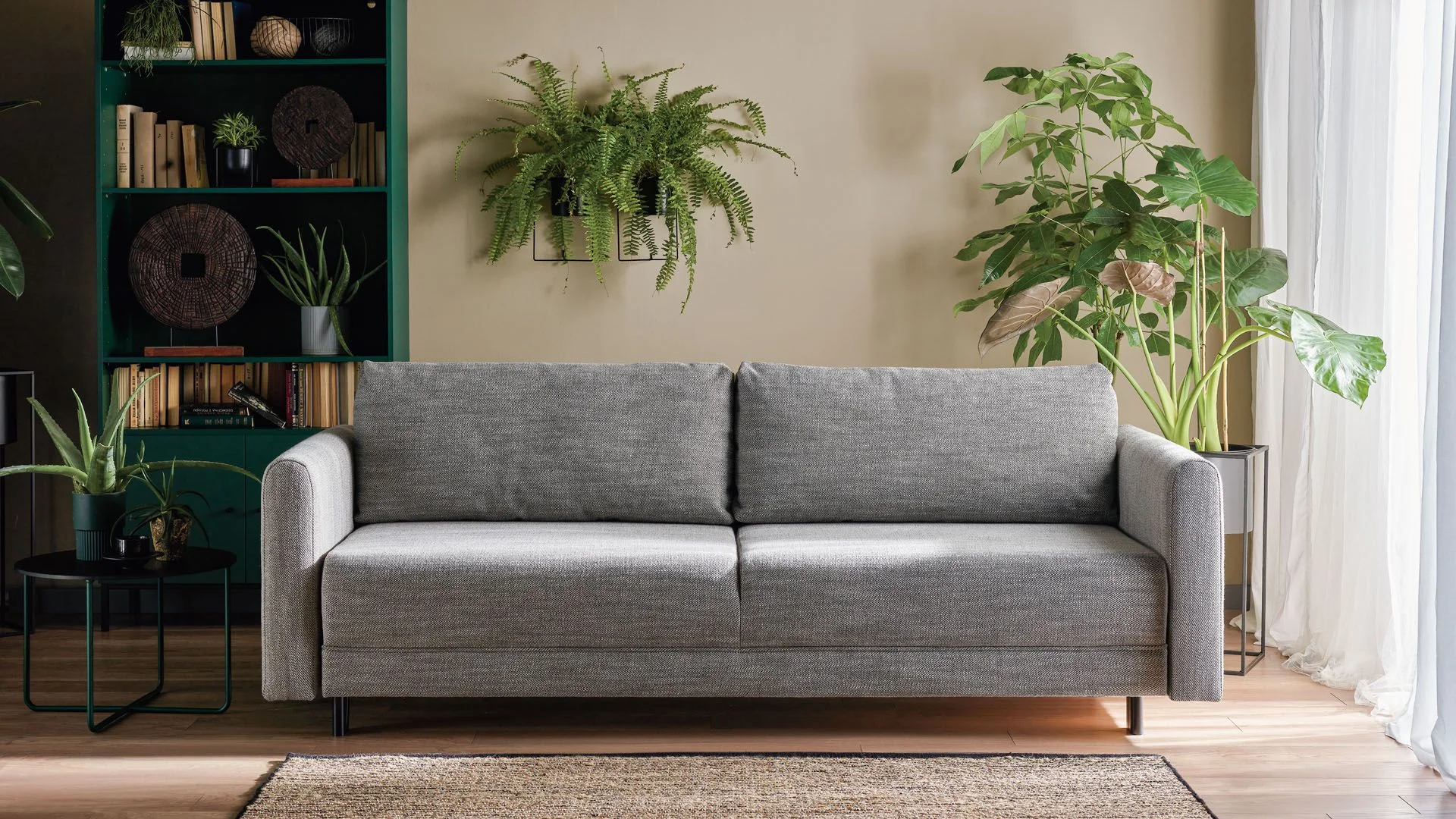 Szara sofa w otoczeniu zielonych roślin i regału z książkami.